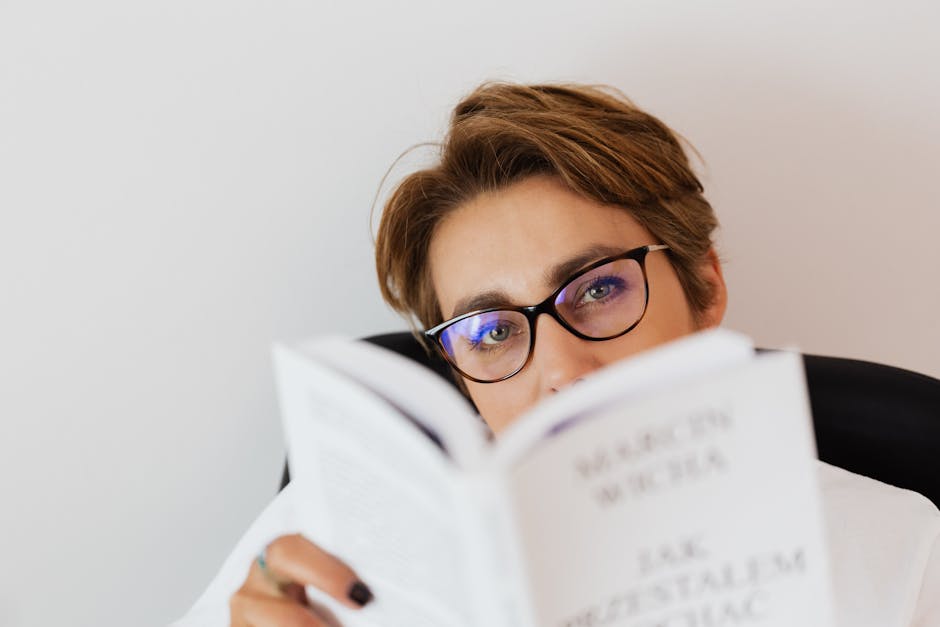 Young female wearing eyeglasses sitting on armchair and reading book while spending free time at home during weekend and looking at camera