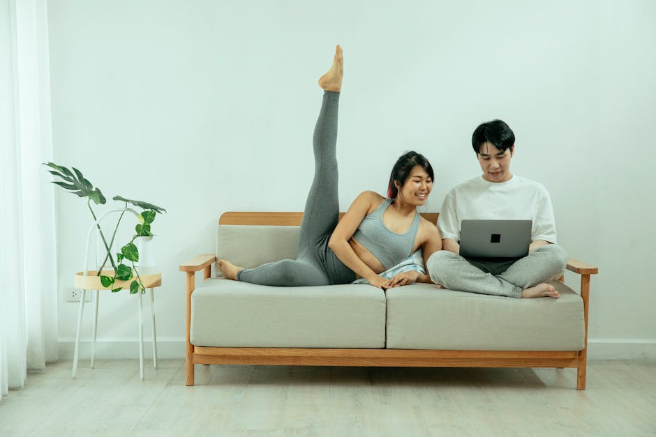 Couple enjoying leisure time on sofa, one using laptop and the other stretching in activewear. Indoors, peaceful atmosphere.